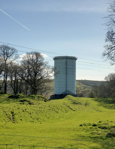 A tall, round water tower in a lush green spring field.  An underground tunnel beneath the grass, leads up to it. 