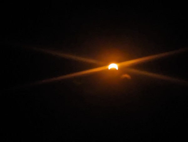 A photo of a crescent sun in the middle of a partial (for me) eclipse. The disc of the moon is at the bottom, and lens flare crisscrosses the sun.