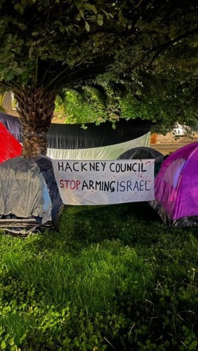 Several tents with a large Palestine flag hung between the trees.  In front of it is a sign reading HACKNEY COUNCIL STOP ARMING ISRAEL
