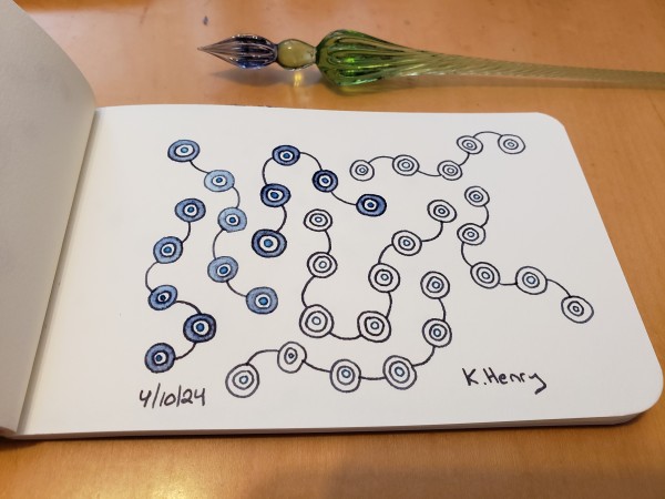 Hand drawn generative art in ink on an open page of my sketchbook. The abstract pattern is a bit like beads on short curvy strings. My glass dipping pen is next to my sketchbook.