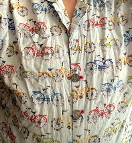 Wrinkled cotton print of classic upright bikes with chainguards, fenders, and racks. They're red, green, blue, and yellow on a light blue background. The buttons are large and metal with an intricate bas-relief of a 19th century cyclist posed next to a bike.