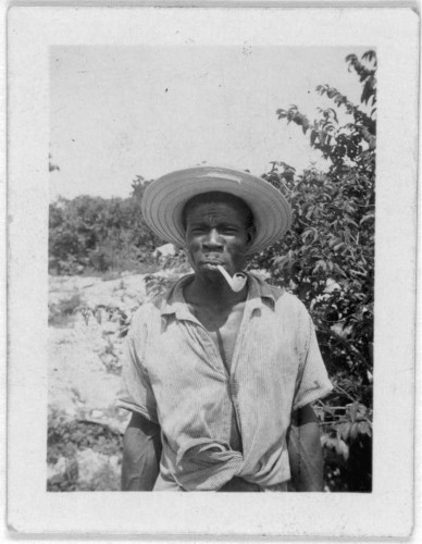  The image is a vintage black and white photograph. It features a man standing in the foreground, wearing a wide-brimmed straw hat and what appears to be a work shirt with rolled up sleeves, suggesting he may have been engaged in manual labor. He has a pipe in his mouth and is looking directly at the camera, exuding a sense of calm or possibly deep thought.

The background shows an outdoor setting with lush greenery and trees, indicating that the photo was taken in a natural environment, perhaps a farm or a rural area. The quality and coloration of the image suggest it may be from an earlier time period, which could be due to its age or the intentional aesthetic choice made by the photographer.

There are no texts visible on the image. However, there is information available about the visit by Alan Lomax and Mary Elizabeth Barnicle to Andros Island in the Bahamas, where this photo might have been taken based on the style of clothing and the landscape features.