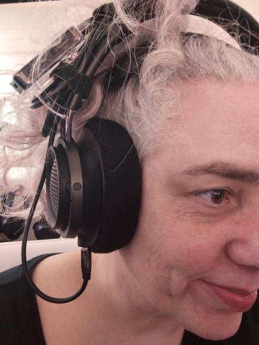 A woman with curly gray hair wearing analog headphones connected to a Bluetooth receiver clip with the cable and clip attached to the headband with a velcro strip. 