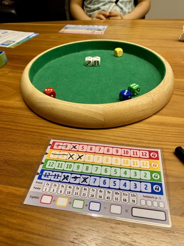 Qwixx-Zettel laminiert mit non-permanent Markern und ein Würfelteller mit bunten Würfeln auf einem Tisch zwischen 2 Spielern. 