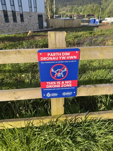 A bilingual "No Drone Zone" sign attached to a wooden fence with grass in the foreground and a building with visible hills in the background.