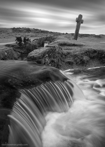 A monochrome photograph capturing a tranquil scene in Devon, England. In the image, a timeless stone monument known as Windy Post Cross stands against the moody sky of Dartmoor. Below, a gentle stream flows, its motion captured in a soft blur, adding a sense of serenity to the rugged landscape.