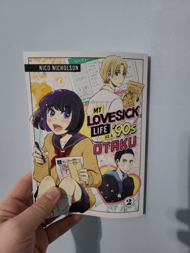 Photograph of a white person's hand holding Volume 2 of My Lovesick Life as a '90s Otaku against a white door. The cover has a teenage girl with dark hair and dark blue eyes, smiling while holding a checklist and an envelope in hand while wearing a Japanese school uniform. In the upper right-hand corner is a picture of a light haired teenage boy's head / shoulders holding paper with a picture of the same girl in his hand. In the lower right-hand corner is a picture of a teenage boy with slicked back dark hair inside of a heart.