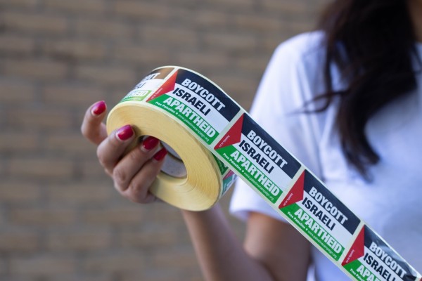 A person with long dark hair & wearing white t-shirt is holding a roll of ‘Boycott Israeli Apartheid’ stickers with the Palestinian flag as the design backdrop.