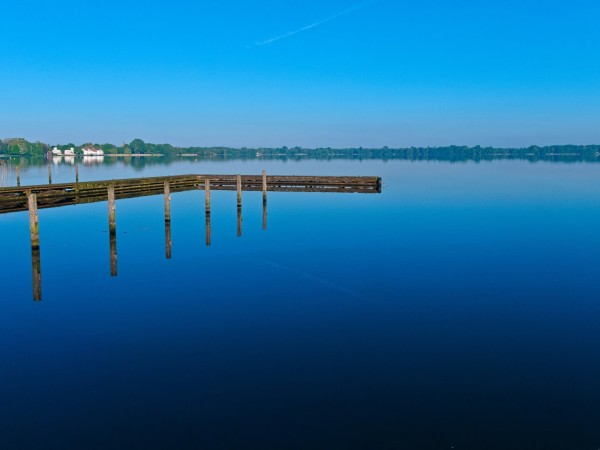 A blue expanse of water. A jetty can be seen on the left, with some wooden piles in front of it. They look long, but their lower part is only a reflection in the water.
A wooded bank and a white building complex can be seen in the background.