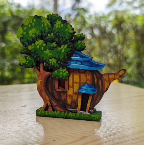 Painted wood cutout of a teapot-shaped house next to a big tree. It has a blue roof, a yellowish facade and a cat sitting in the window. 3 layers of wood make it a bit 3D