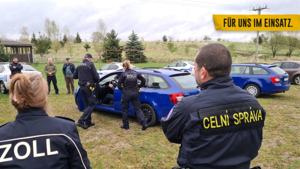 Im Vordergrund stehen eine deutsche Zöllnerin und ein tschechischer Zöllner mit dem Rücken zur Kamera. Vor ihnen übt ein deutsches Zollteam eine Pkw-Kontrolle an einem blauen Fahrzeug.