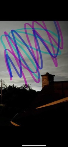 Image description: A photo of a dark evening sky with scattered clouds, featuring blue and pink digital scribbles across it. The bottom of the photo shows silhouettes of rooftops and trees.