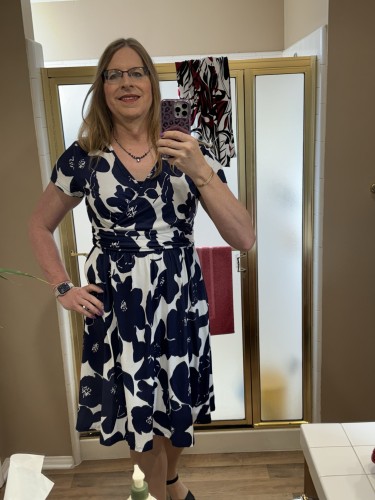 A woman taking a mirror selfie in a bathroom, wearing glasses, a blue and white floral dress, and holding a phone with a patterned case.