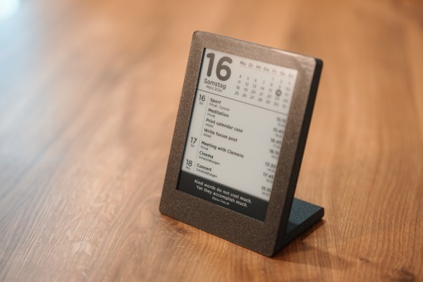A photo of my epaper calendar standing on a desk. It is enclosed in a 3d printed housing and the display shows a calendar with all the days of the month and a list of appointments below.