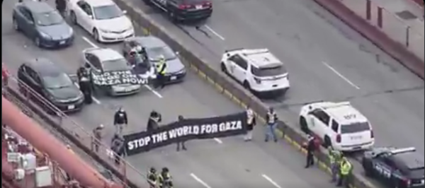 A small group stretching across the roadway, holding banners reading STOP THE WORLD FOR GAZA and END THE SIEGE ON GAZA NOW