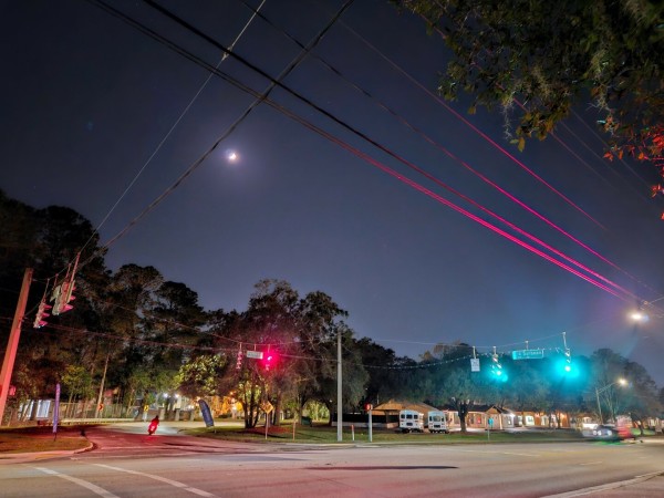 Beneath a night sky with the glow of a partial moon a street intersection glows red and green from hanging traffic signals, reflecting their colors upon nearby power lines. A motorcycle is seen in silhouette having just passed through the intersection, it's tail light glowing red.