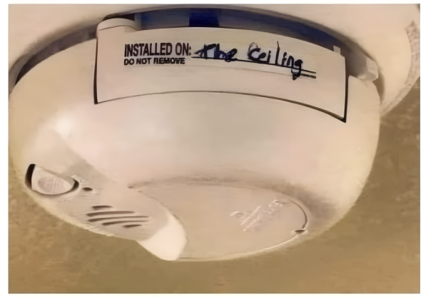 Photo of a smoke detector haning on the ceiling. On its side is a label reading "Installed on." Instead of the date, someone wrote "the ceiling" next to that.