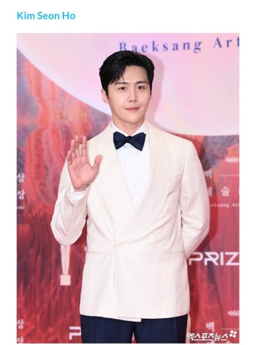 Actor Kim in a white suit with a bow tie waving at a red carpet event with a stage backdrop.