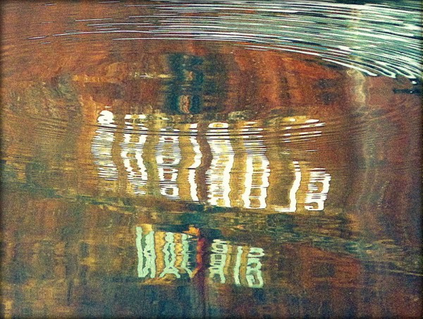 A colour photograph showing the stern of a small wooden boat reflected in water. The name, Glorious and home port, Girvan are prominent in white, fragmented by the motion of the water against a natural wood background and concentric ripples top of frame.