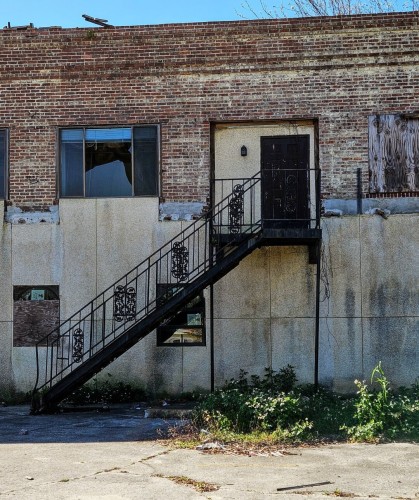 From a desolate parking lot with weeds, dirt build up, and trash scattered, a view directly at an old two story brick building, long vacant and in disrepair.  Windows boarded up.  One door, at the top of a old black metal stairs is also sealed up. One of many in the area on this major downtown corridor.