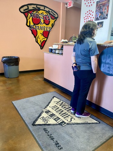 A woman standing at the counter, ordering pizza. She stands on rug that reads
Angie’s & Jimmie’s
Pizzeria
EST. 1985
602-246-7833

On the wall is another rug with the company name sans apostrophes. The rug on the wall is shaped like a slice of pizza. 