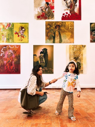 A woman crouched next to a standing child in an art gallery, with various paintings on the walls. The child appears to be talking or pointing, and the woman is looking at the child while holding her hand.