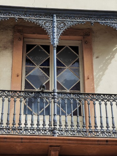 A balcony with a geometrically divided double casement window behind.

The balcony rail and scalloped decoration to the eaves are created from fine filigree  wrought iron, in details evoking lilies, daisies and dolphins