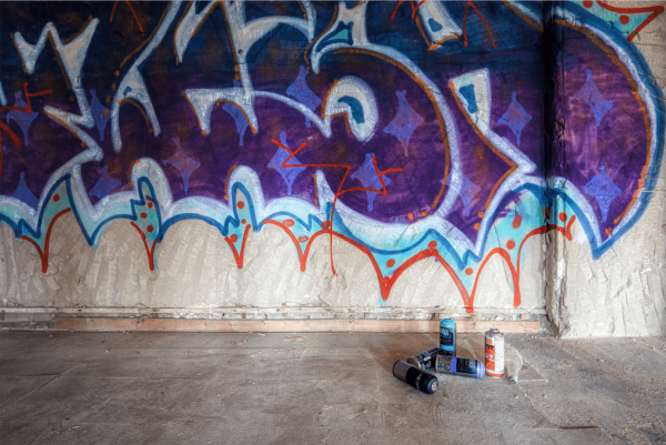 The image shows an indoor scene with a section of a wall covered in graffiti. The graffiti art is composed of vibrant colors with predominant hues of purple, blue, and red, featuring abstract patterns and shapes. There appears to be a symbol or letter-like shapes, but they don’t form recognizable words. The texture of the wall suggests that the paint layers may be thick and possibly still fresh, as indicated by the glossiness in some areas.

On the floor, there's a collection of used spray paint cans in blue, black, and orange, lying scattered, with one can standing upright. The floor is concrete, with a worn, dusty surface suggesting an abandoned or seldom-used building. Natural light enters from the left side, creating a subtle lighting gradient across the scene, and emphasizing the texture of the wall and floor. This setup suggests that the photograph might have been taken during or shortly after the creation of the graffiti.