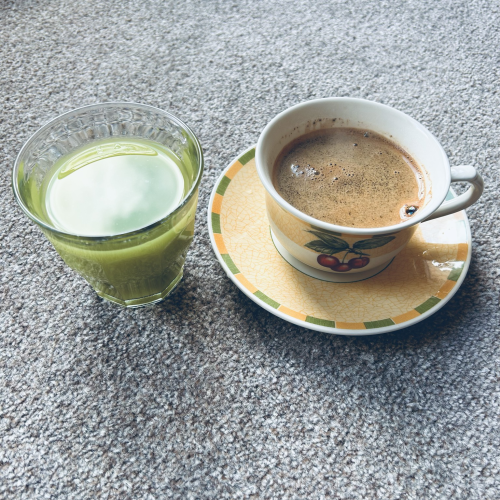 A square photo in colour. Two morning beverages sit beside each other, one cold, one hot. To the left is a small clear Duralex glass of green juice, to its right is a steaming cup of black coffee in a white & yellow cup that sits on a matching saucer.