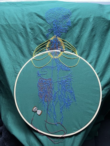An image of an old engraving of a vagus nerve is embroidered on green cotton. The gut is in a large bamboo embroidery hoop. The embroidery of the main nerve lines is done in blue chain stitch in perlé cotton. The brain and some of the face is stitched in the same thread in backstitch. Details in the face are stitched in single strand floss. A bright red anatomical heart is stitched in red whipped back stitch. Around it a bright golden yellow halo in a vesica piscis shape is stitched in single strand thread. Three sets of ribs have are stitched in bright green/yellow stem stitch. There are some tiny details in different colours along some of the nerves. Some burgundy roiling is being stitched in the left gut area. A needle minder in the shape of an hourglass that reads, "This is taking forever," sits near the stitches holding a needle.