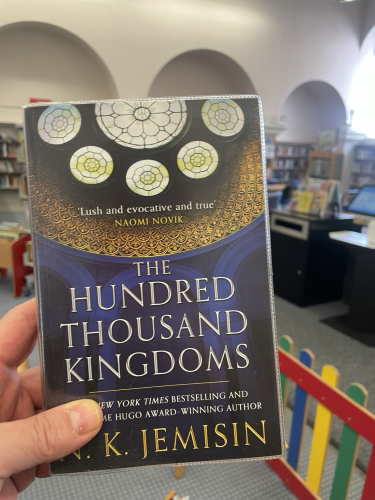 A copy of N.K. Jemisin’s ‘The Hundred Thousand Kingdoms’ held in front of a library scene 