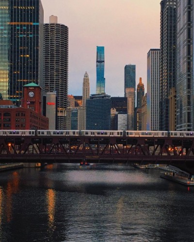 A subway crosses the Chicago River at golden hour. Multiple skyscrapers are in the background, in soft focus. 