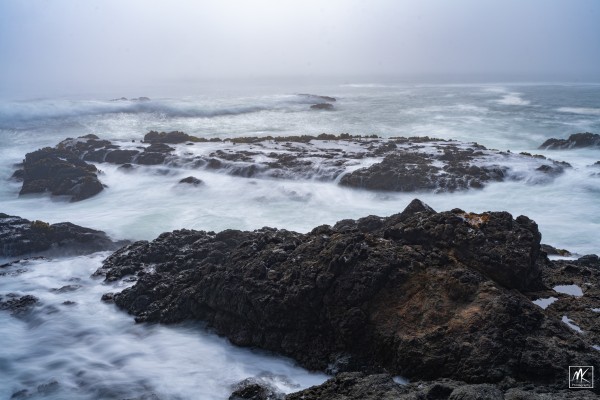 Color photo of a coastal seascape with water and waves flowing over low rocks and foggy skies above. 