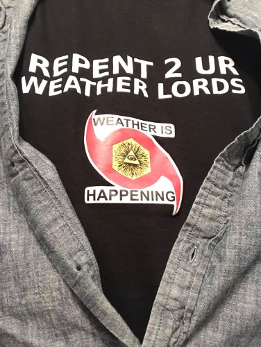 A weather is happening tee shirt saying repent 2 ur weather lords and the weather is happening logo, which is a hurricane symbol with the illuminati pyramid eye in the center. it's a black tee shirt viewed through an opened dark blue linen button shirt