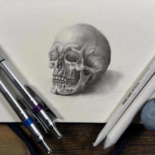 Pencil-drawn skull with shading on paper, flanked by mechanical pencils and eraser.&10;