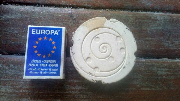 A plaster disc with a spiral pattern on a wooden table, and a matchbox for size comparison