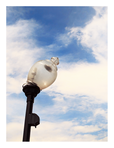 close-up. midday sun. a translucent, plastic acorn-style streetlight globe leans right, atop a black metal pole, also leaning right. the background is large unformed clouds in a baby blue sky.