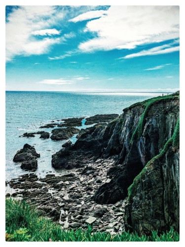 Color photo showing a view of the Celtic Sea taken from a cliff in County Cork, Ireland. There are green-topped cliffs on the right, a rocky beach below, and white clouds in a pale blue sky over the horizon.