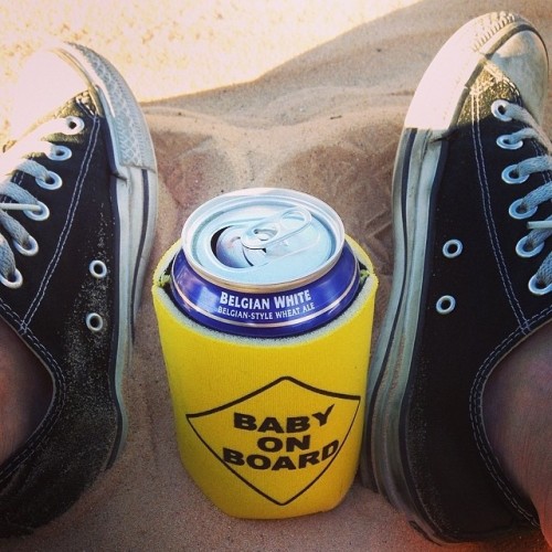 Yellow beer koozie that says "baby on board" in black letters. It's on a can of blue moon in some sand on a beach, all in between two black Converse All Stars
