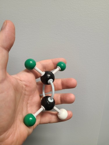Organic chemistry molecular model kit in a white person's hand. The model has two black balls with angled connections between them. The top black ball has two green balls connected to them. The bottom one has a single white ball and one green ball attached.