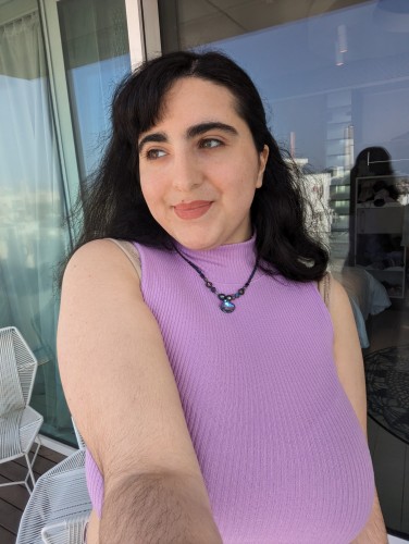 photo of me in pink top, looking to the side with a small smile
I am modelling a handmade beaded necklace with a stunning soft lilac purple - white triangle glass pendant
I have pink lipstick and green eyes
dark brown hair
