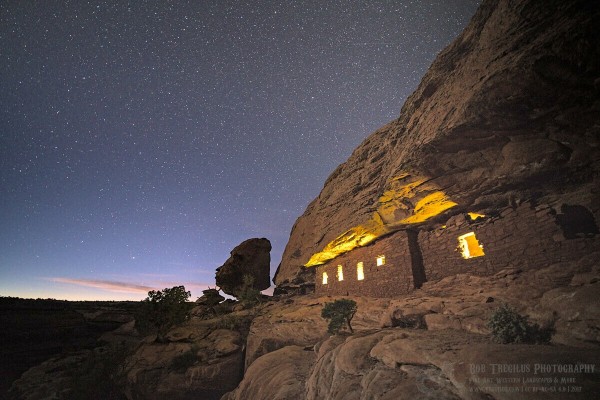 A photo showing a cliff-dwelling after sunset. The brick like structure fills a large crack in a sandstone cliff on the right side of the frame. There are five rectangular windows each with a yellow glow coming out as if there were a fire inside. A couple of small bushes grow on the surrounding rock. At the far end, silhouetted against the darking sky, is a large balanced rock the looks like a human head in profile. Some stars are visible in the sky and the last glow of evening is on the horizon.