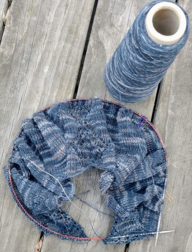 The beginning of a knitting project on needles, with a cone of yarn standing next to it, laid out on a weathered wooden floor.

Yarn is in shades of indigo blue - from very washed out to very dark, giving the piece a variegated look.

Knitting is mostly stockinette with a stripe of lace in the center and along the sides.