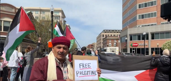 Small group of protesters with huge Palestinian flags.  An Asian gentleman in a red hat is centre foreground, holding a home made cardboard sign reading FREE PALESTINE