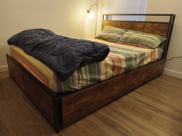 A full-size bed, with a modern bedframe of brown-stained wood and black-painted metal accents, made up with bedlinens in earthtone stripes, and a folded dark blue comforter laid crosswise at its foot. A floor lamp and window brighten the scene.