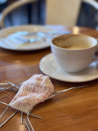 Foreground, in focus, sock knitting in progress. Heel flap and turned heel visible. Background, half drunk latte in a mug, and an empty plate with utensils (crepe already eaten)