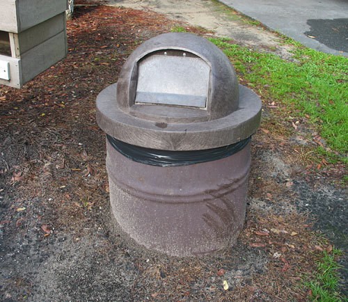 A round trash can buried in the ground near grass. 