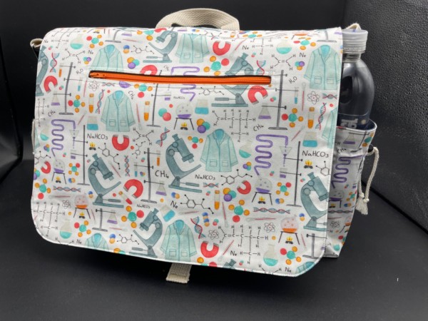 A messenger bag with a white background and brightly colored watercolor-style illustrations of science-themed stuff, like lab coats and microscopes and magnets and beakers with purple stuff in them and molecules and chemical formulas and dna and such. There is an orange zipper in the flap, as well as a side cargo pocket holding a water bottle. 