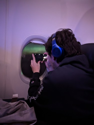 A young man in an airplane window seat taking a picture of the Northern Lights with a cell phone through the airplane window 