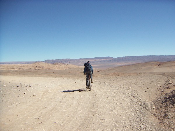 Une personne qui roule à vélo vue de dos, qui a un grand sac à dos et un bonnet. Le chemin qui n'est pas très égal tourne vers la gauche, et à l'horizon sous le ciel bleu des montagnes désertiques s'étendent derrière un plateau.